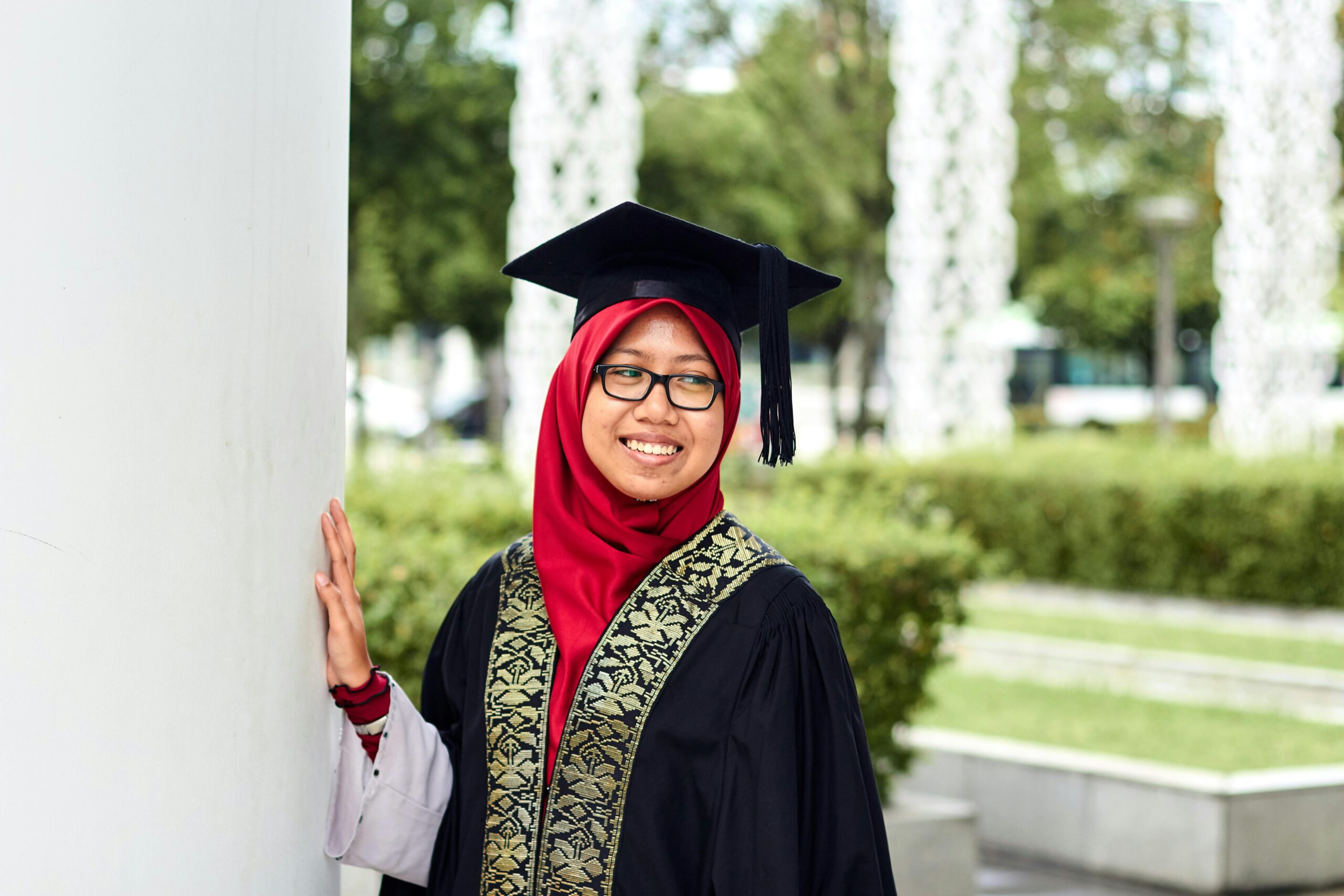 A smiling Southeast Asian woman in cap and gown celebrating graduation outdoors in Putrajaya.